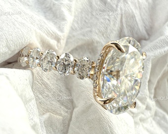 May include: A gold engagement ring featuring a large, oval-cut diamond as the centerpiece, accented by smaller diamonds along the band. The ring is set against a white, textured background, showcasing its brilliance and design.
