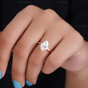 May include: A gold ring featuring a pear-shaped, clear gemstone. The ring is set on a thin gold band. The ring is shown on a person's hand with blue nail polish.