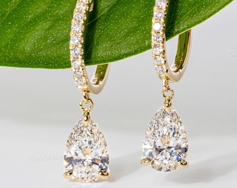 May include: A pair of gold hoop earrings with a row of small, clear stones along the top. Each earring has a pear-shaped, clear gemstone pendant. The earrings are displayed against a white background.