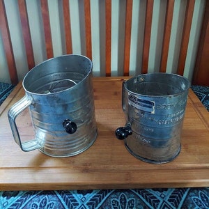 May include: Two vintage metal flour sifters with black handles. The sifters have a silver finish and are sitting on a wooden surface.