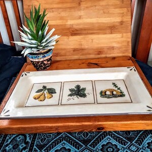 May include: A white rectangular serving tray with three compartments. Each compartment features a different design: pears, a leaf, and a bridge. The tray is on a wooden surface with a potted succulent plant.