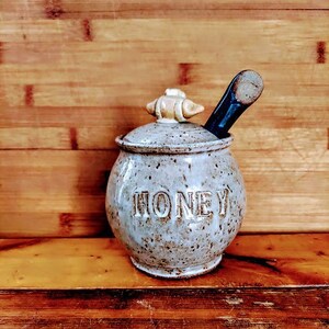 May include: A white ceramic honey pot with a brown lid and a wooden honey dipper. The pot has the word "HONEY" written on the front.