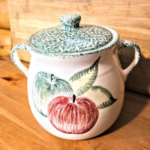 May include: A white ceramic casserole dish with a lid, featuring a green speckled pattern. The dish has two handles and is decorated with hand-painted apples in red and green, with green leaves.