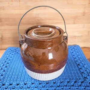 May include: A brown and white glazed ceramic crock with a metal handle. The crock has a white band around the bottom and a brown lid.