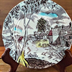 May include: A vintage ceramic plate with a brown and white illustration of a quaint village scene. The plate features a church, houses, trees, and people walking down a path. The plate has a blue and white rim.