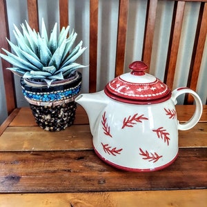 May include: White teapot with red and white trim and a red leaf design. The teapot is sitting on a wooden surface next to a potted succulent.