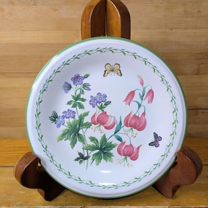 May include: A white ceramic plate with a green rim and floral design. The plate features pink and purple flowers, green leaves, and butterflies. The plate is displayed on a wooden stand.