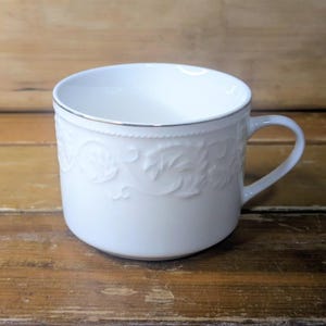 May include: White ceramic teacup with a silver rim and embossed floral design. The cup has a handle and sits on a wooden surface. The teacup is a classic design for serving tea or coffee.
