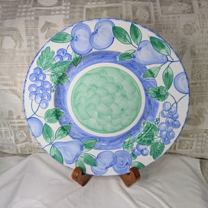 May include: A large, round, hand-painted ceramic plate with a fruit design. The plate features blue apples, pears, and grapes with green leaves on a white background. The center is green, surrounded by a blue border.