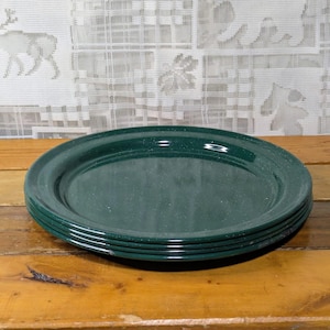 May include: A stack of five dark green enamel plates with a speckled finish. The plates are oval-shaped and have a slightly raised rim. They are arranged on a wooden surface, with a patterned white curtain in the background.