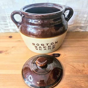 May include: A brown and beige ceramic bean pot with a lid. The pot has two handles and the words "BOSTON BAKED BEANS" are printed on the front. The lid is round with a small knob.