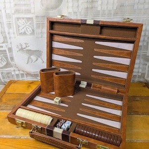 May include: Open brown leather backgammon set with dice, game pieces, and two leather dice cups. The game board has a brown and white design. The set is on a wooden surface.