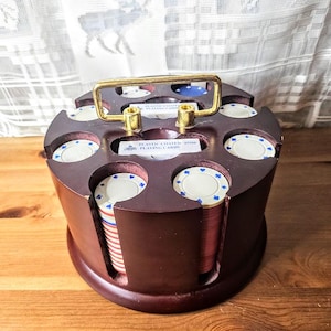 May include: A wooden poker chip holder with a gold handle, filled with stacks of red, white, and blue poker chips. The holder has slots for chips and a small card holder. The text on the card holder reads "PLASTIC COATED 97556 PLAYING CARDS."