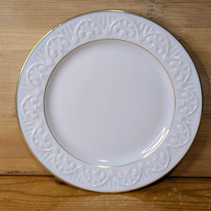May include: A white porcelain plate with a gold rim and a decorative embossed floral pattern around the edge. The plate is set against a wooden background, showcasing its elegant design.