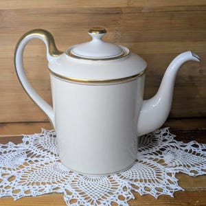 May include: A white porcelain teapot with a gold handle, rim, and lid accents. The teapot is cylindrical with a curved spout and sits on a white, lacy doily. The background is a wooden surface.