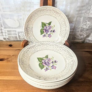 May include: A stack of vintage white plates with a floral violet design. The plates have a gold filigree border and a central image of purple violets with green leaves. The plates are stacked on a wooden plate stand.