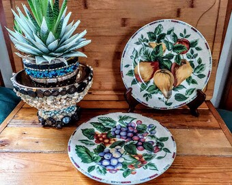 May include: Two decorative plates with a fruit and floral design. The plates are on a wooden surface with a potted succulent plant.