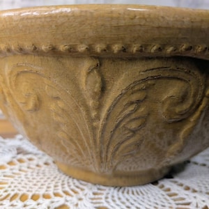 May include: A tan-colored ceramic bowl with embossed floral patterns. The bowl has a decorative beaded rim and a wide, rounded shape. The bowl is sitting on a white lace doily.