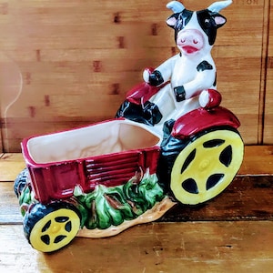 May include: A ceramic planter shaped like a red tractor with a black and white cow figurine driving it. The planter has a removable top and a yellow wheel.
