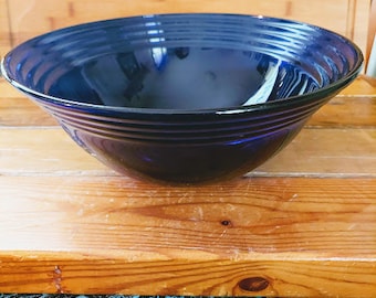 May include: A deep blue ceramic bowl with a ridged exterior. The bowl is set on a wooden surface with a patterned blue and black fabric in the foreground. The bowl's interior reflects the light.