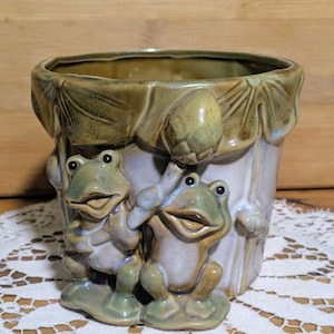 May include: A ceramic planter with a whimsical design featuring two green frogs. The frogs are sculpted in a playful pose, adding a touch of charm. The planter has a light green and white color scheme, with a textured surface.