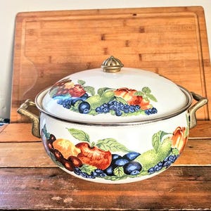 May include: White enamel Dutch oven with a fruit design. The design features apples, pears, grapes, and blueberries. The lid has a brass knob.