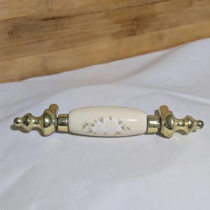 May include: Cream-colored cabinet pull with a floral design and gold-colored hardware. The center section features a white rose and green leaves. The hardware has a decorative, rounded design.