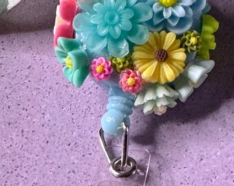 May include: A blue and white floral badge reel with a pink flower charm. The reel is made of plastic and has a metal clip.