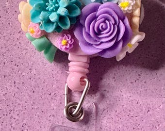 May include: A pink retractable badge holder with a floral design. The flowers are made of polymer clay and include a purple rose, blue and white daisies, and a teal succulent. The badge holder has a clear plastic strap and a teal daisy charm.