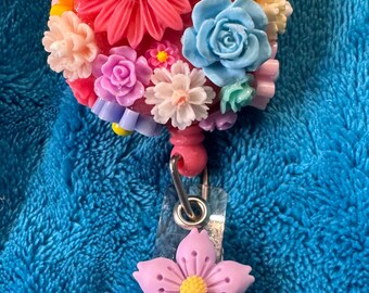 May include: A colourful flower-shaped badge holder with a pink, blue, yellow, and white flower design. The badge holder has a clear plastic strap and a pink flower charm.