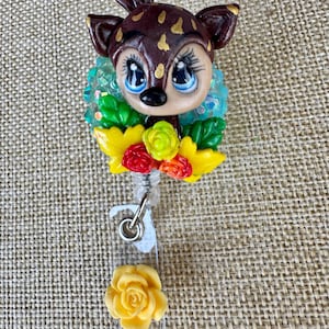 May include: A handcrafted badge reel featuring a deer with brown fur and gold spots, blue eyes, and a floral collar. The reel includes a yellow rose at the bottom. The badge reel is designed for use with an ID badge.