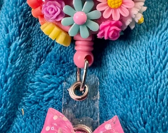 May include: A pink and blue floral badge reel with a pink polka dot bow. The reel has a clear plastic holder and a metal clip.