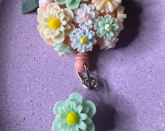 May include: A pink and green floral badge reel with a clear plastic badge holder. The reel features a cluster of pastel pink, green, and white flowers.