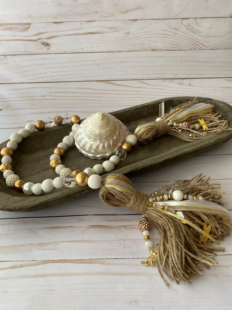 May include: A white and gold beaded garland with a burlap tassel and a white seashell. The garland is displayed on a wooden tray.