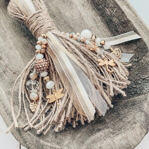 May include: A beige and gold tassel with beads, a dragonfly charm, and a heart charm that says "Made with Love".