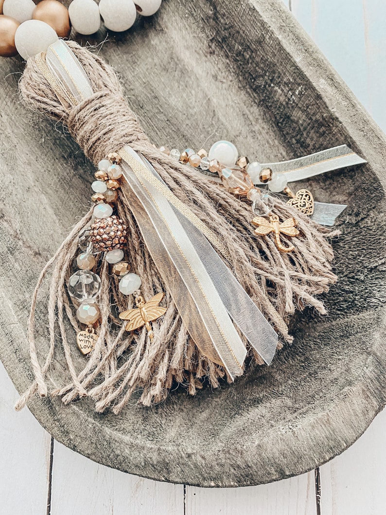 May include: A beige and gold tassel with beads, a dragonfly charm, and a heart charm that says "Made with Love".
