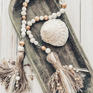 May include: A white and gold beaded garland with brown tassels hangs in a wooden bowl. The garland features white and gold beads, and the tassels are made of brown jute. A seashell is also in the bowl.