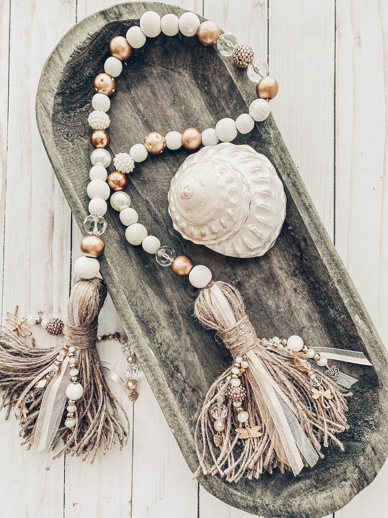 May include: A white and gold beaded garland with brown tassels hangs in a wooden bowl. The garland features white and gold beads, and the tassels are made of brown jute. A seashell is also in the bowl.