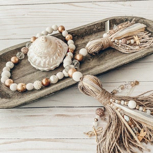 May include: A wooden bowl filled with a beaded garland featuring white, brown, and gold beads. The garland has a large, white seashell in the center. Two brown and gold tassels hang from the bowl.