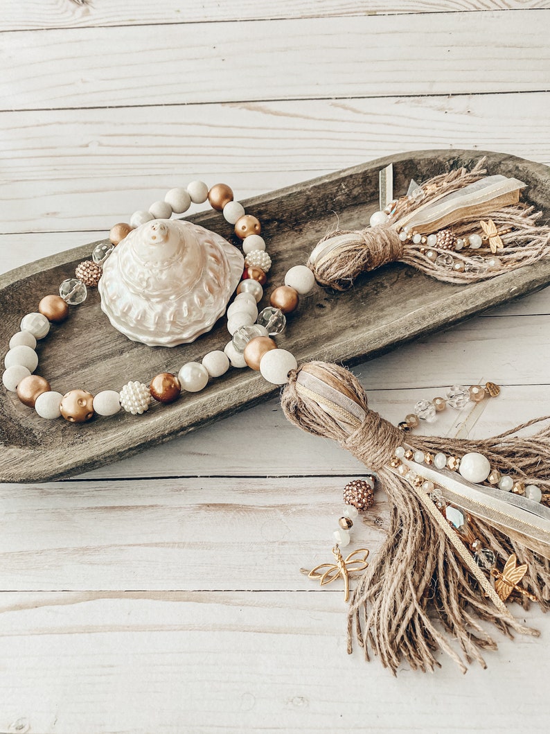 May include: A wooden bowl filled with a beaded garland featuring white, brown, and gold beads. The garland has a large, white seashell in the center. Two brown and gold tassels hang from the bowl.