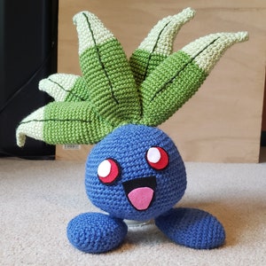 May include: A blue and green crocheted plush toy with a smiling face and red eyes. The toy has a leafy green head and a blue body with small arms and legs.