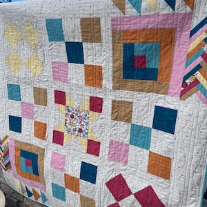 May include: A handmade quilt with a cream background and colourful patchwork squares. The quilt features squares in shades of pink, teal, brown, and orange, with a floral center. The quilt is hanging outside with a blue sky in the background.