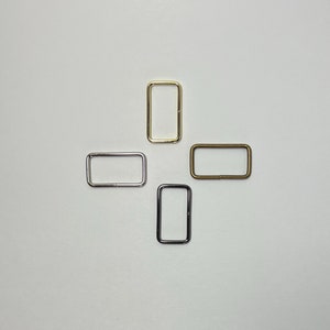 Puede incluir: Cuatro anillos metálicos rectangulares en oro, plata, bronce y negro dispuestos sobre una superficie blanca. Estos componentes de hardware se utilizan en varios proyectos de artesanía y costura.