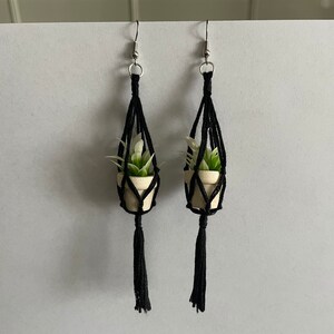 May include: A pair of black macrame earrings with a small white pot and a green plant hanging from each earring.