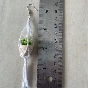 K&ouml;nnte beinhalten: Ein wei&szlig;er Makramee-Pflanzenh&auml;nger mit einer kleinen Sukkulente im Topf. Der H&auml;nger hat einen wei&szlig;en Quasten und ist etwa 5 cm lang.