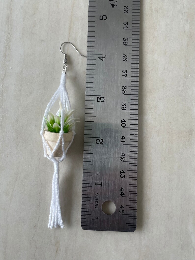 K&ouml;nnte beinhalten: Ein wei&szlig;er Makramee-Pflanzenh&auml;nger mit einer kleinen Sukkulente im Topf. Der H&auml;nger hat einen wei&szlig;en Quasten und ist etwa 5 cm lang.