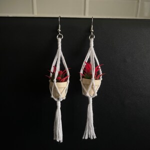 K&ouml;nnte beinhalten: Ein Paar Ohrringe mit einer kleinen Topfpflanze, die an einem wei&szlig;en Makrameemuster h&auml;ngt. Die Pflanzen haben rote Bl&auml;tter und stehen in wei&szlig;en T&ouml;pfen.