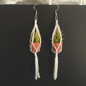 May include: A pair of white macrame earrings with small potted plants hanging from them. The plants have green leaves and are in terracotta pots.