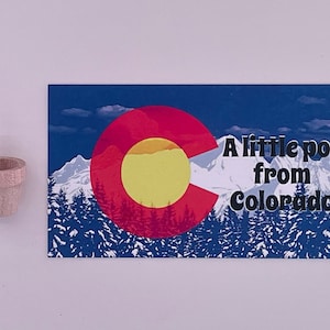 May include: A small, unfinished wooden pot next to a rectangular print. The print features the Colorado state flag design, a mountain scene, and the text "A little pot from Colorado."
