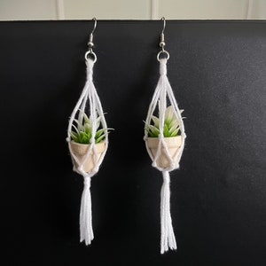 May include: A pair of white macrame earrings with a small potted succulent hanging from each earring. The earrings have a white tassel hanging from the bottom.
